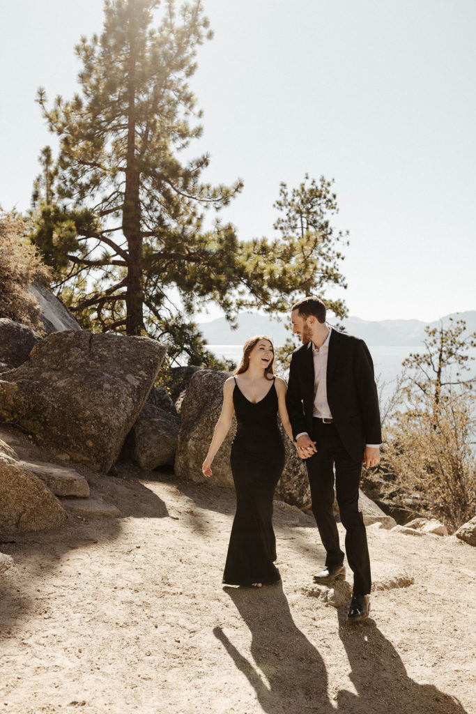 Engagement couple holding hands while walking along dirt trail and smiling at each other at Lake Tahoe