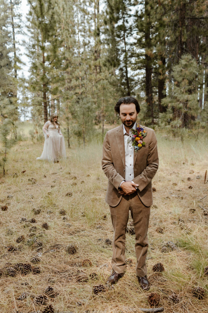 Wedding groom waiting while bride walks up behind him for first look in forest in Lake Tahoe