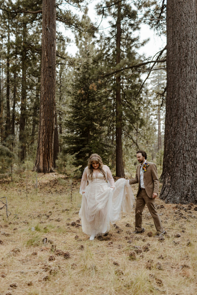 Wedding groom carrying brides dress tail as they walk through the forest in Lake Tahoe