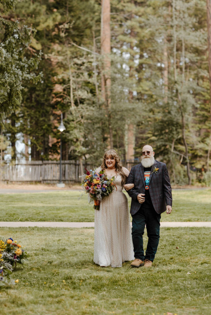 Wedding bride walking down wedding aisle while holding dads arm and smiling outside in Lake Tahoe