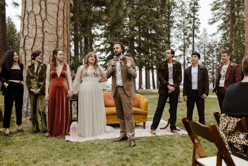 Wedding groom holding microphone and giving speech while bride looks at him with wedding party in background outside in Lake Tahoe