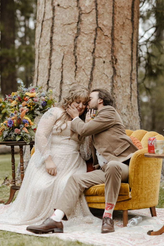 Wedding couple sitting on a couch together while groom kisses brides cheek outside in Lake Tahoe