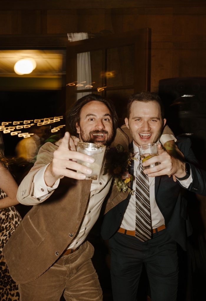 Wedding groom and groomsman holding drinks while pointing at camera  as lights blur behind them in Lake Tahoe