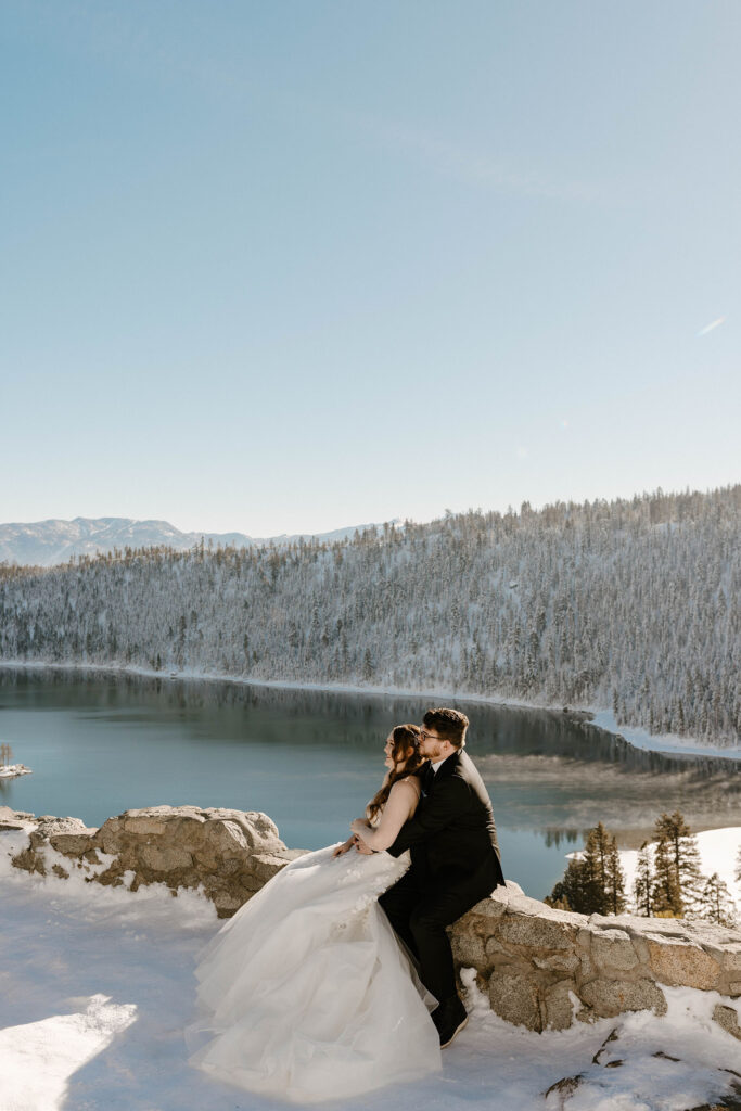bride and groom sitting on a ledge smiling and hugging in lake tahoe from afar