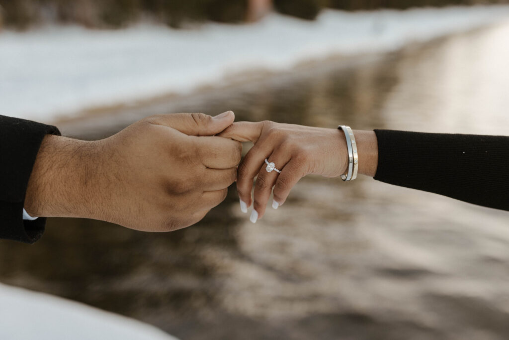man and woman holding hands and showing off engagement ring overlooking the snow and water in lake tahoe