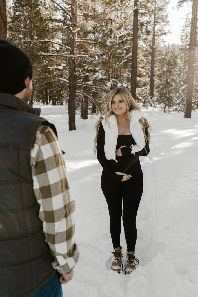 pregnant woman holding belly and smiling at her husband in the snow in lake tahoe