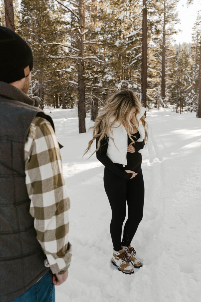 pregnant woman holding belly in the distance while her husband watches her in lake tahoe