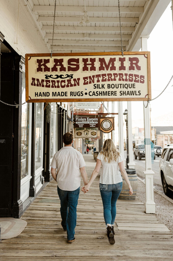 Engagement couple holding hands and walking down wooden walkway with wooden signs overhead at virginia city
