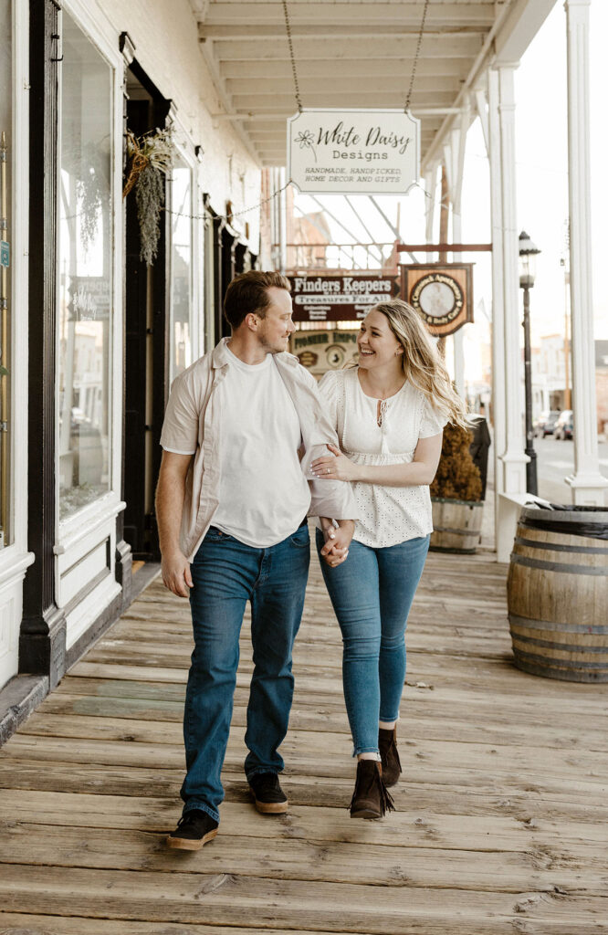 Engagement couple holding hands and smiling at each other while walking down wooden walkway in virginia city