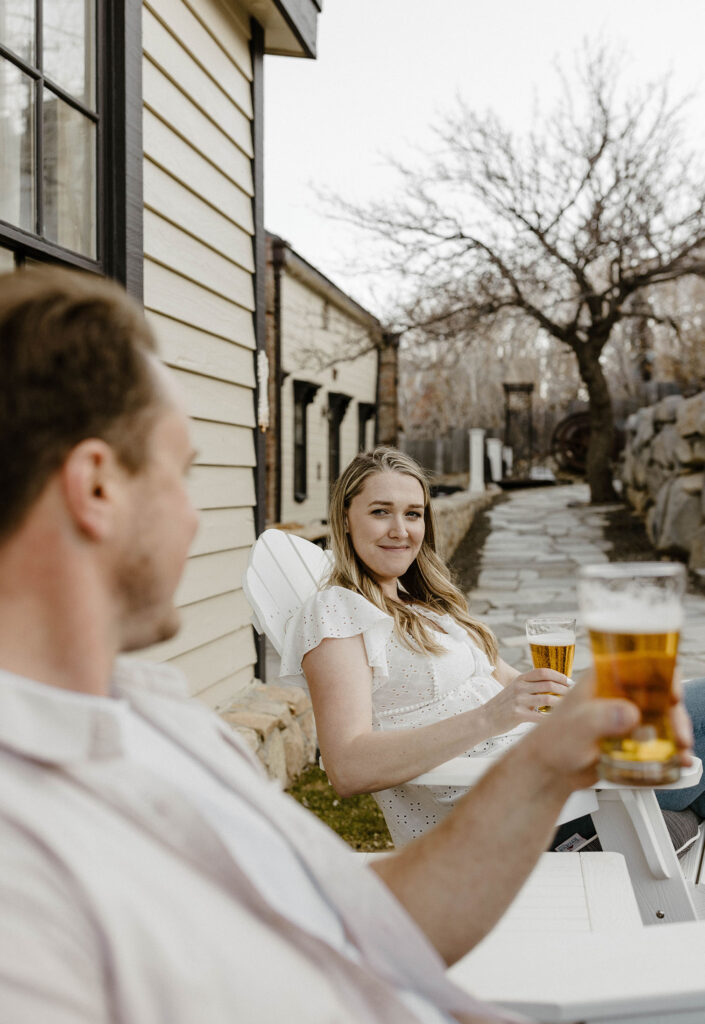 Woman cheering fiancé with beer glass while sitting in white wooden chairs in virginia city