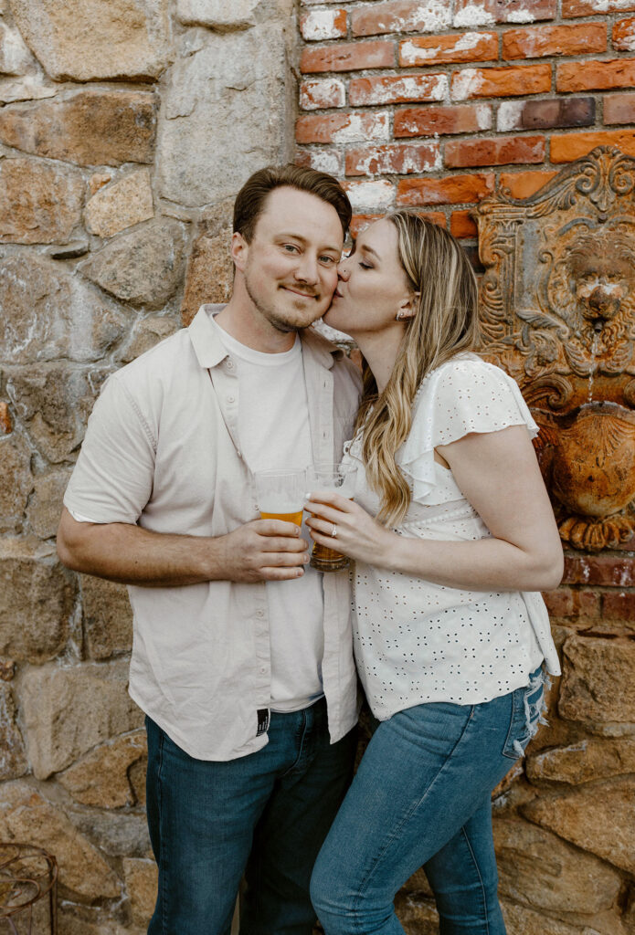 Woman kissing fiancé on cheek while leaning against stone wall in virginia city