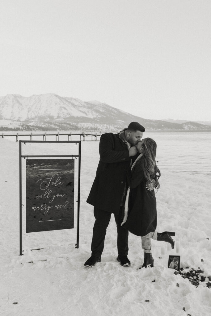 man and woman kissing standing in front of a sign asking the woman to marry him in lake tahoe