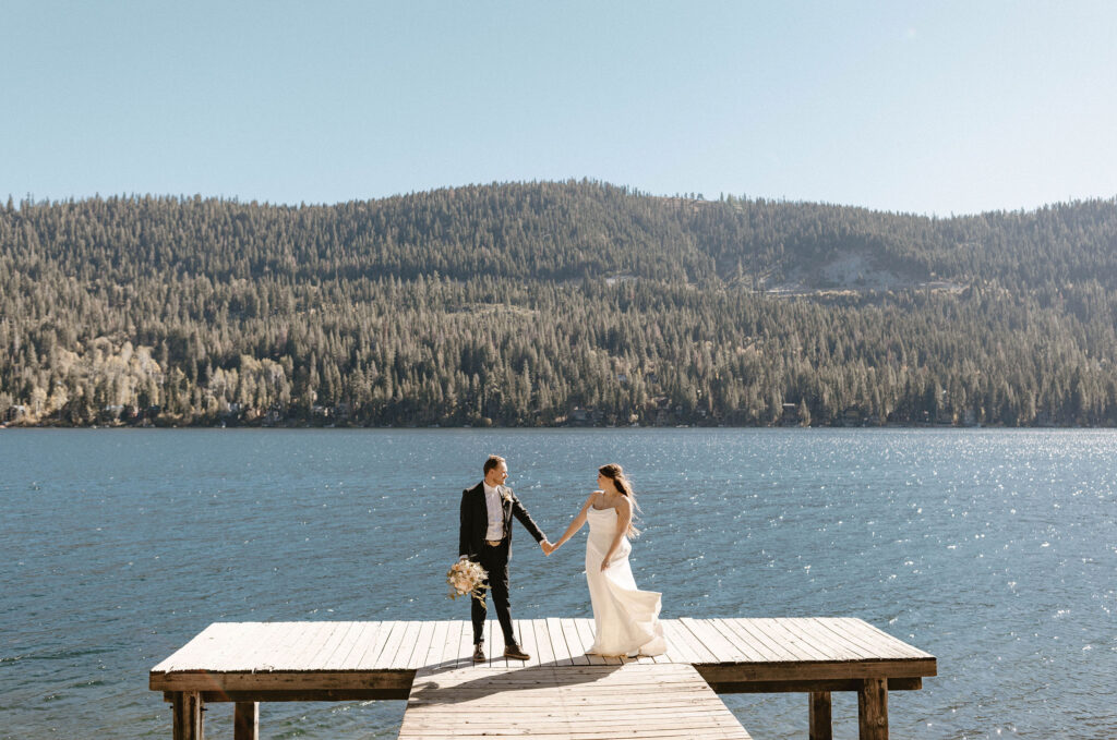 Elopement couple holding hands while looking at each other and standing on dock at Donner lake together with mountains in background
