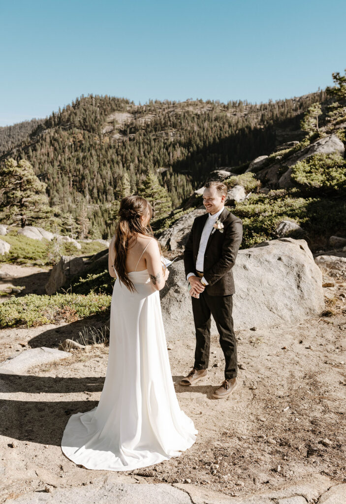 Husband smiling at wife while reading vows with trees and mountain in background at Donner lake