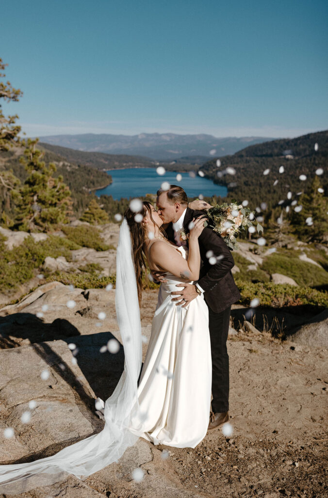 Elopement couple kissing while flower petals fly around them with Donner lake and mountains in background