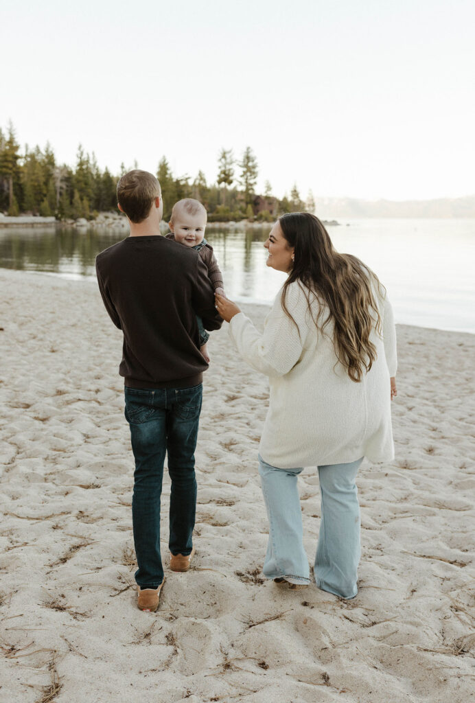 Dad carrying baby while baby smiles at camera and mom plays with baby on beach in Lake Tahoe