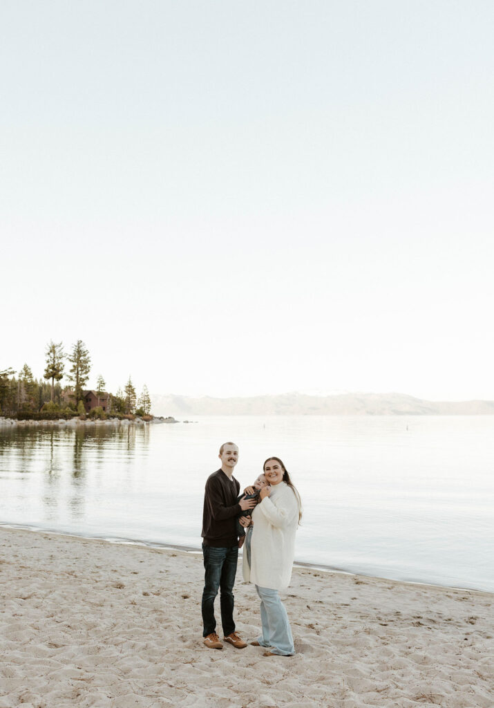 Engagement couple holding baby while smiling at camera while on a sandy beach in Lake Tahoe