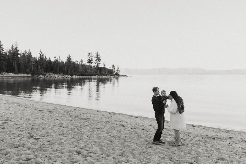 Engagement couple holding and playing with baby while standing on sandy beach in Lake Tahoe