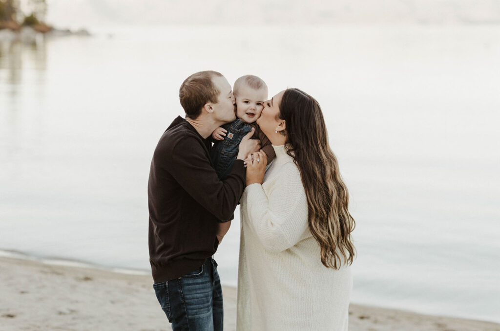Engagement couple kissing baby on either side of face while baby smiles at camera on a beach in Lake Tahoe