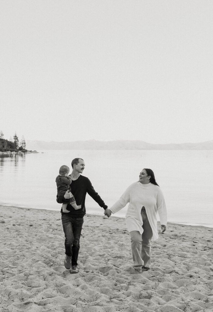 Engagement couple holding hands while walking along sandy beach in Lake tahoe while dad carries baby