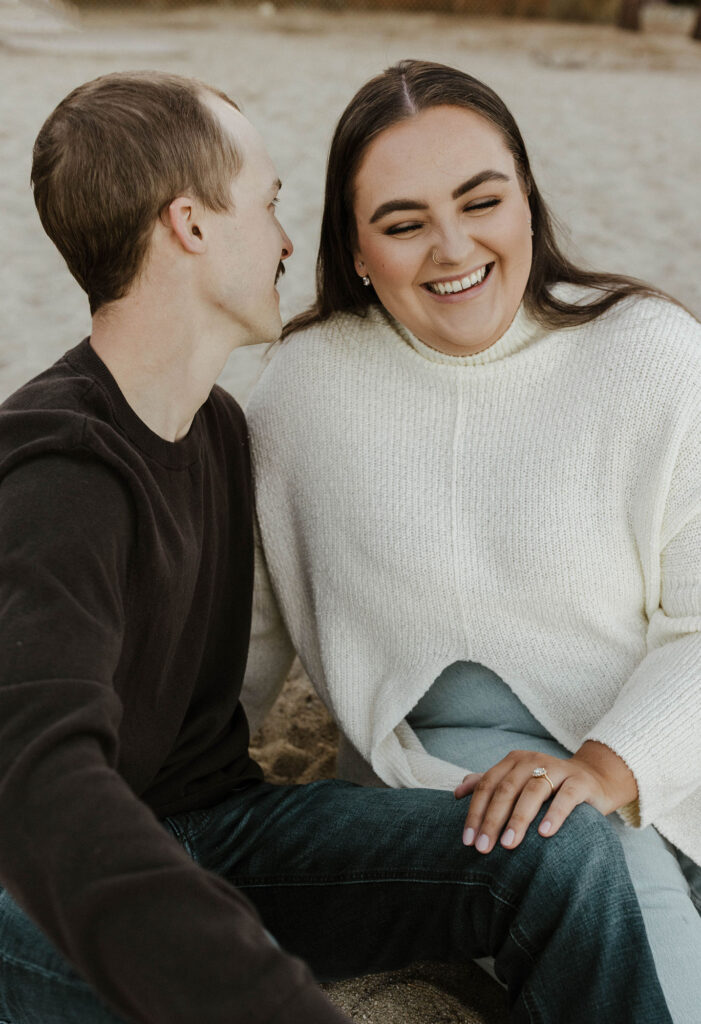 Woman with hand on fiancé's leg smiling while sitting on a sandy beach in Lake Tahoe