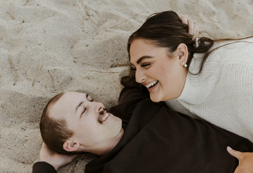 Engagement couple lying on sandy beach holding and smiling at each other in Lake Tahoe
