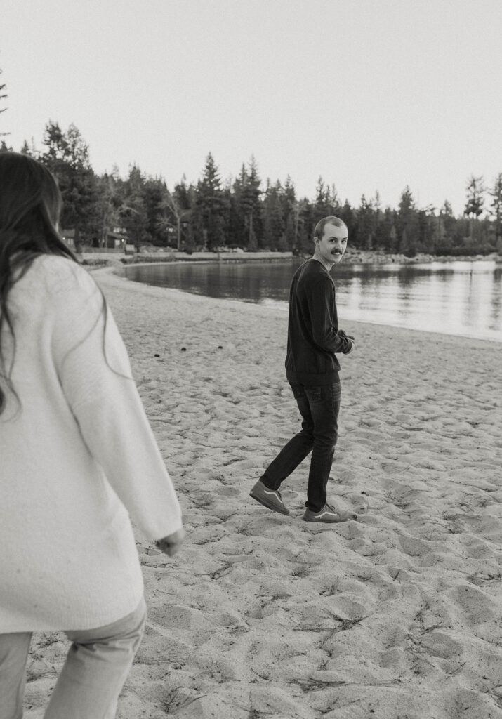 Man looking back over shoulder as fiancé chases him across sandy beach in Lake Tahoe