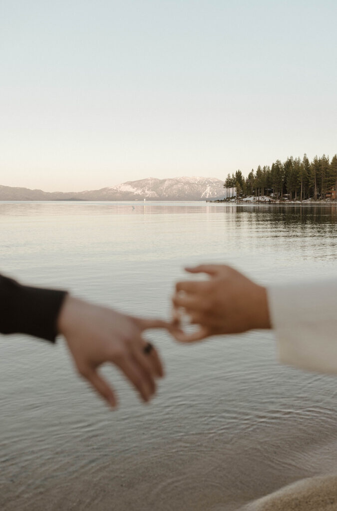Engagement couple holding hands with engagement rings while in Lake Tahoe with mountains and Lake in background