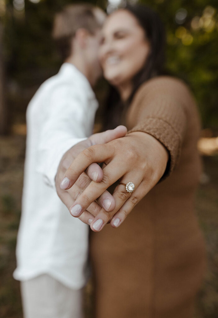 Close up of engagement couple's hands with engagement ring while couple smiles in the background in Lake Tahoe