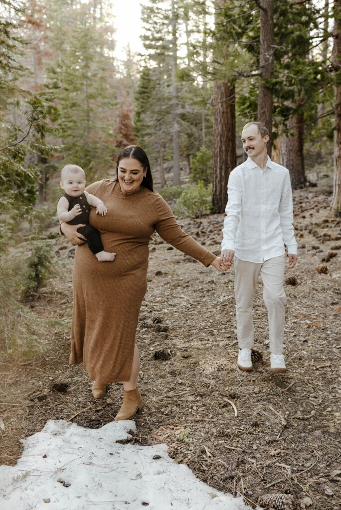Mom carrying baby while engagement couple holds hands and walks through Lake Tahoe forest while baby smiles at camera