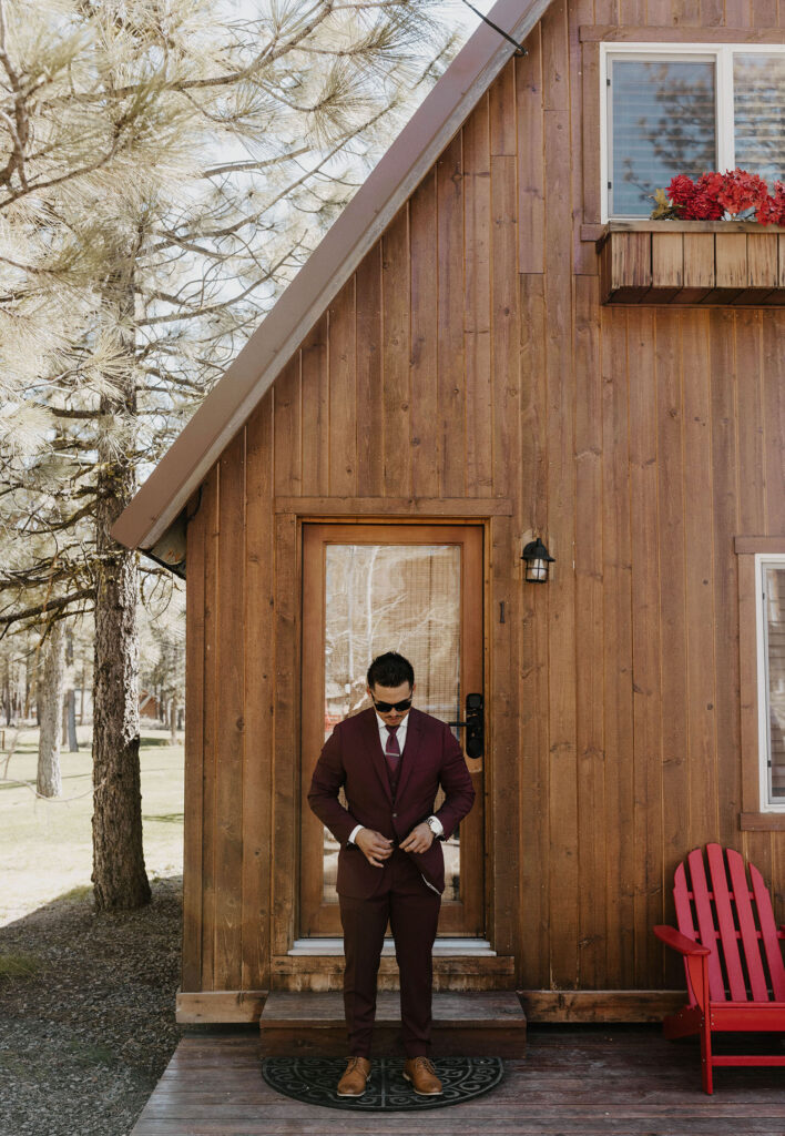 Wedding groom buttoning jacket while standing in front of wooden lodge at Chalet View Lodge
