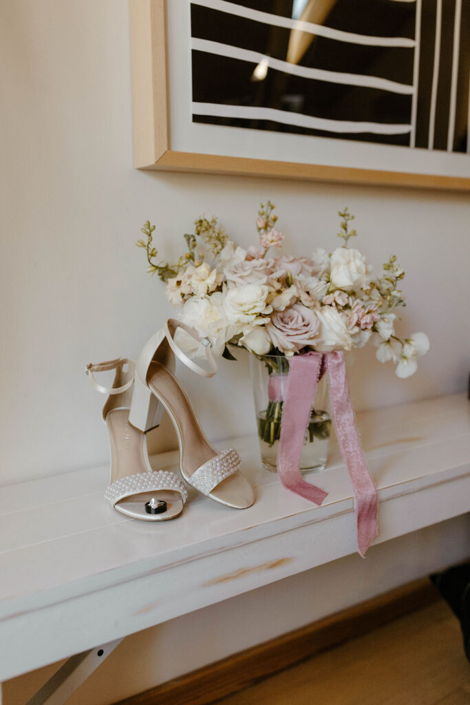 Bride's wedding shoes on white shelf next to floral bouquet with pink ribbon at the Chalet View Lodge