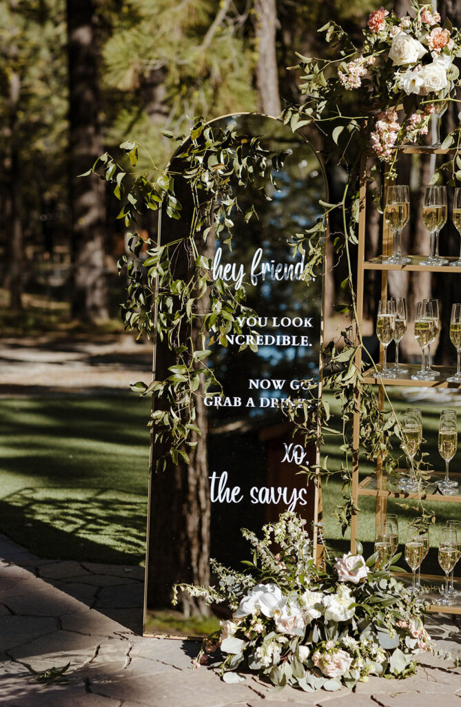 Wedding sign covered in green vegetation and florals with glasses next to it at Chalet View Lodge
