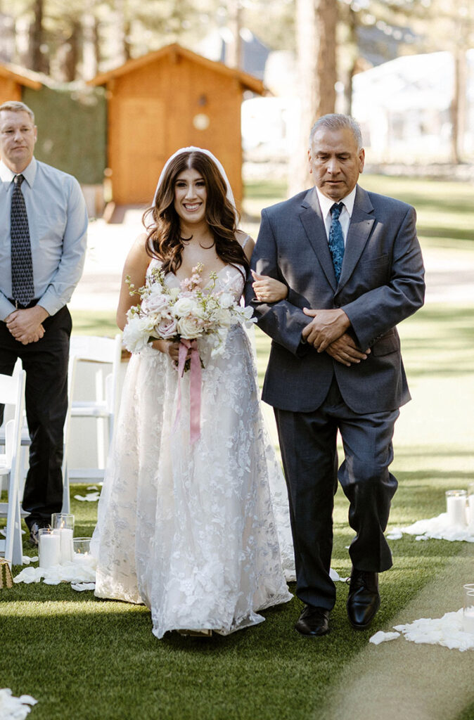 Bride smiling and holding bouquet while being walked down the wedding aisle by dad at Chalet View Lodge