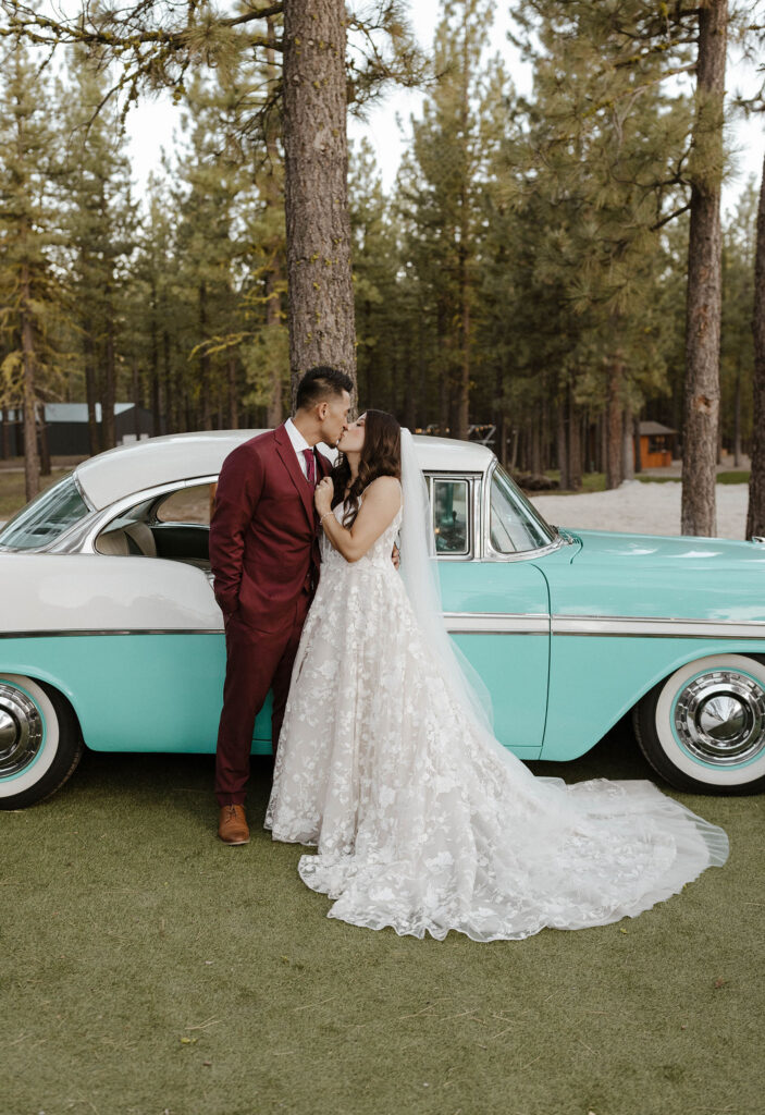 Wedding couple kissing in front of classic teal and white car with tress in background at Chalet View Lodge