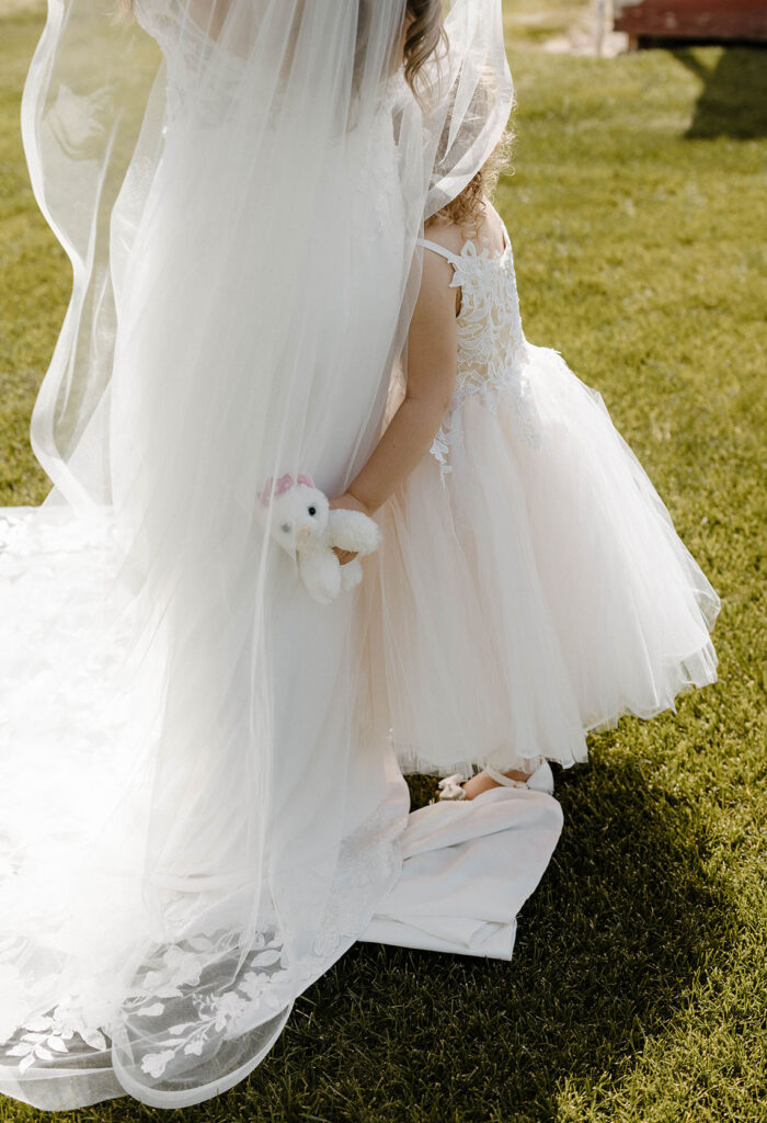 Little girl hugging bride in wedding dress while holding stuffed animal outside at little bear creek ranch