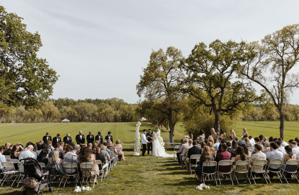 Wedding ceremony with couple holding hands and all guests with wedding arch with white florals at little bear creek ranch