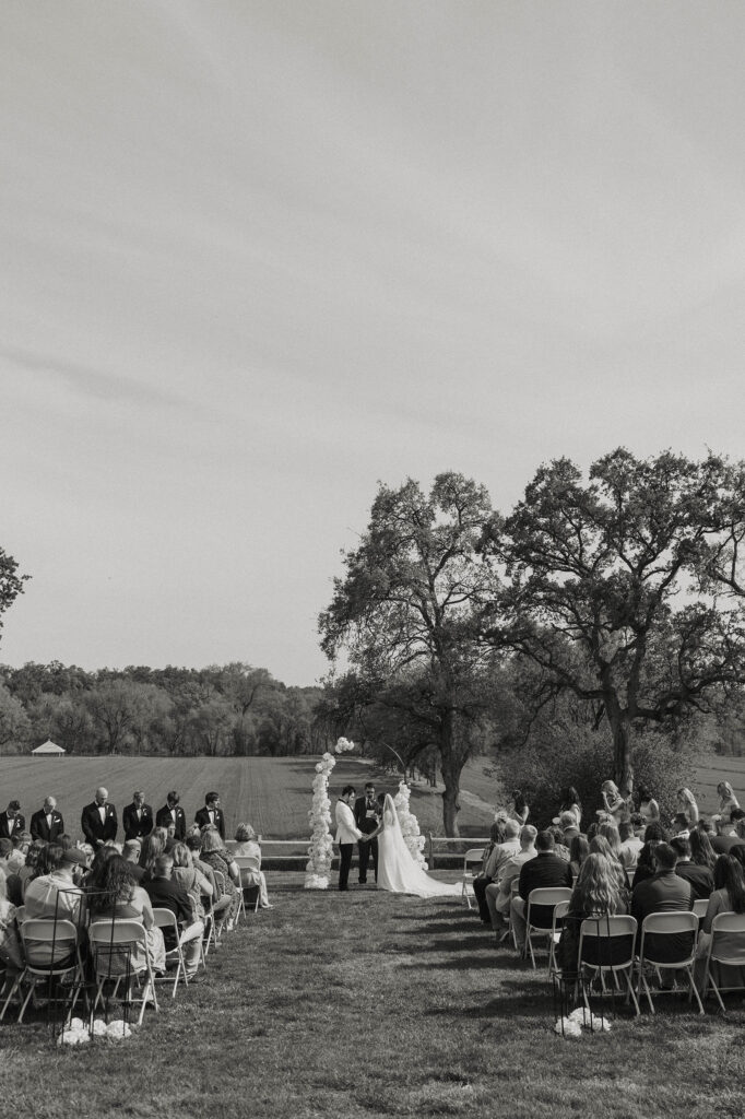 Wedding ceremony with guests and arch with white florals and large field in background at little bear creek ranch
