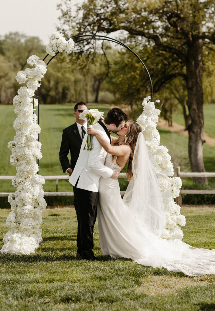 Wedding couple first kiss during ceremony at little bear creek ranch with arch with white florals in background