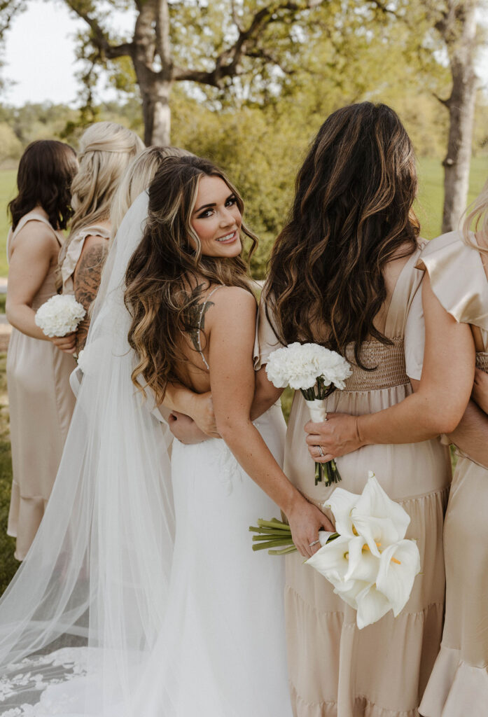 Bride and bridesmaids with arms around each other holding flower bouquets as bride looks back over shoulder at camera at little bear creek ranch