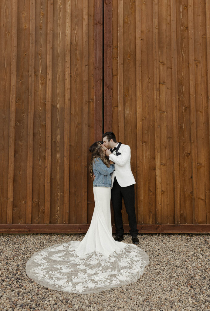 Wedding couple holding and kissing each other while standing in front of large wooden barn doors with gravel at little bear creek ranch