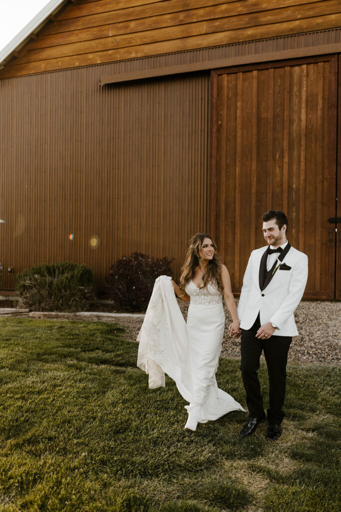 Wedding bride smiling at groom while holding hands and walking together away from brown barn at little bear creek ranch