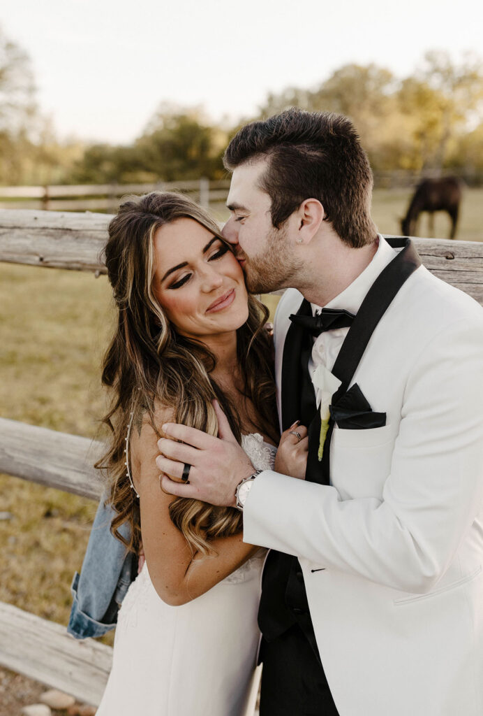 Wedding groom kissing bride on the cheek with wooden fence and horses in background at little bear creek ranch