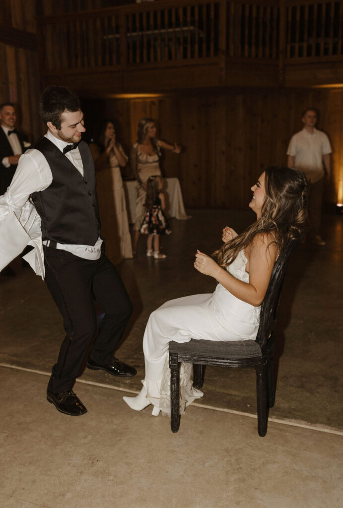 Wedding groom dancing while taking jacket off while bride is sitting on chair laughing during reception at little bear creek ranch