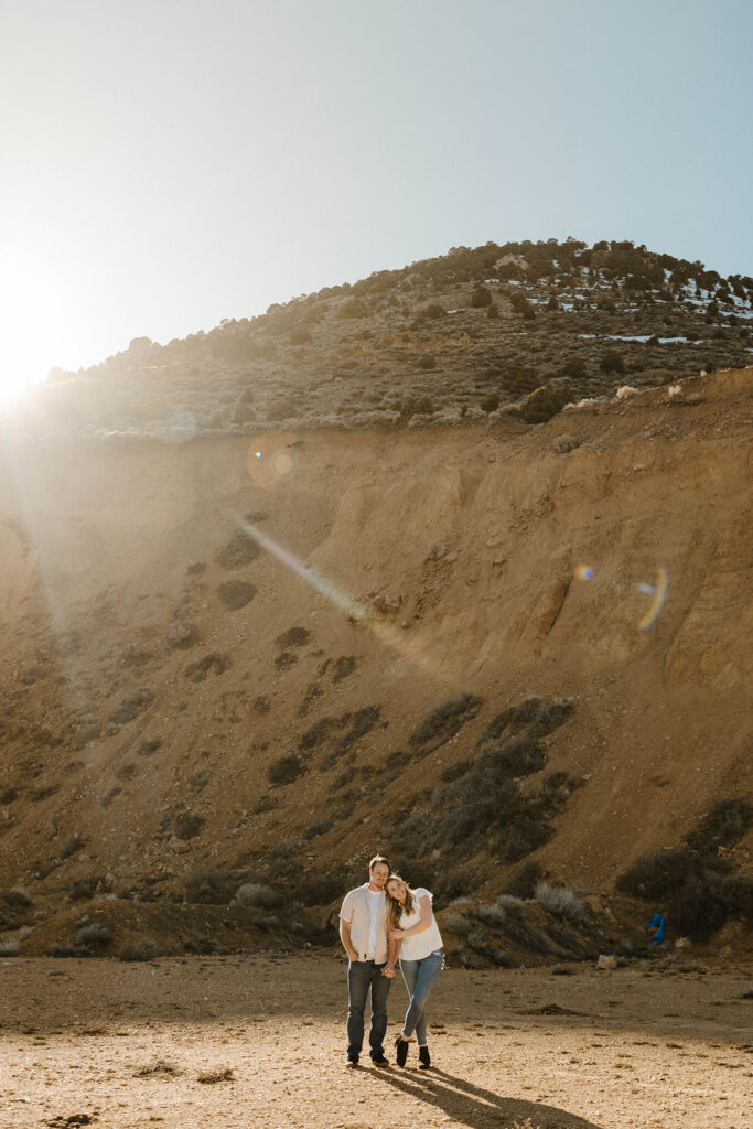 Engagement couple holding hands and leaning on each other while smiling at camera and standing in front of large dirt hill