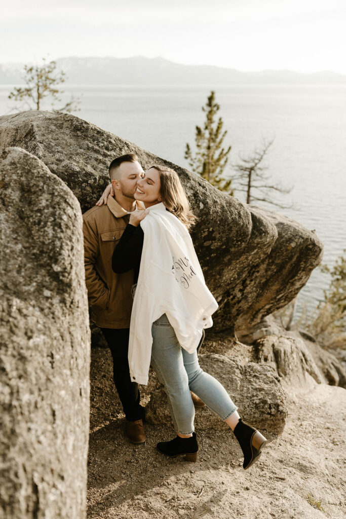 Man kissing fiancé on cheek while leaning on large rock with Lake Tahoe and mountains in background at Logan Shoals