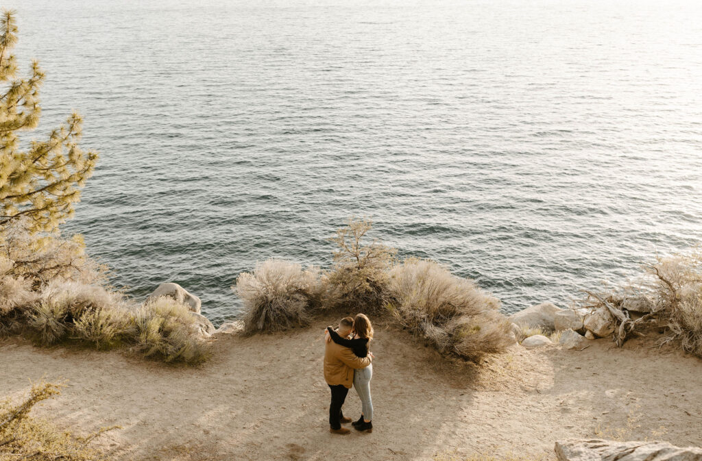 Engagement couple hugging each other while looking at water and standing on dirt path near edge of Lake Tahoe at Logan Shoals