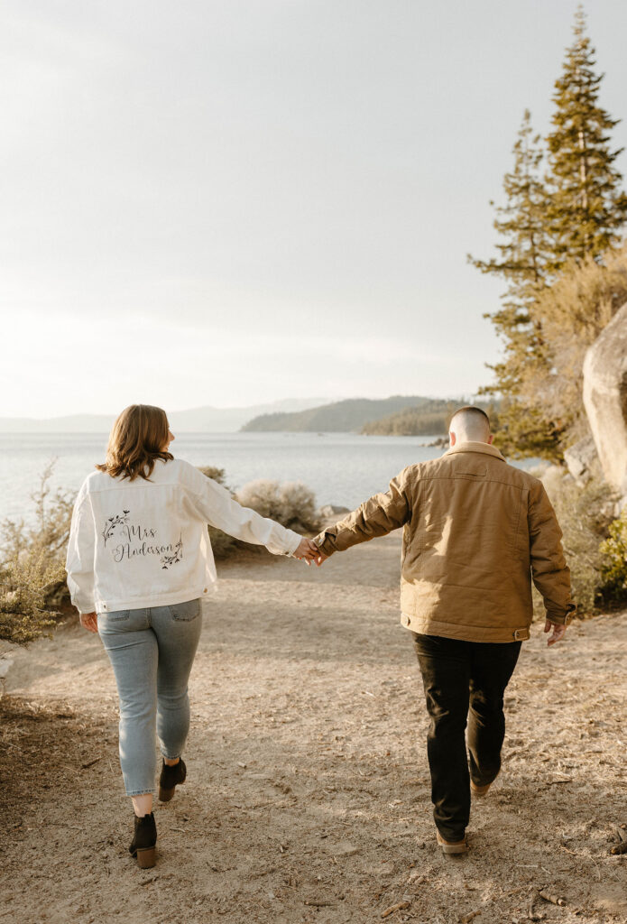 Engagement couple holding hands while walking along dirt trail away from camera while woman is wearing engagement jacket with Lake Tahoe in background