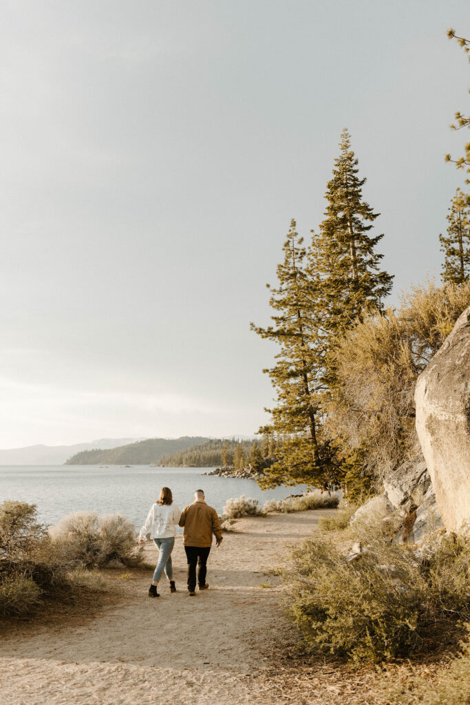 Engagement couple holding hands and walking along dirt trail at Logan Shoals away from camera with Lake Tahoe in background