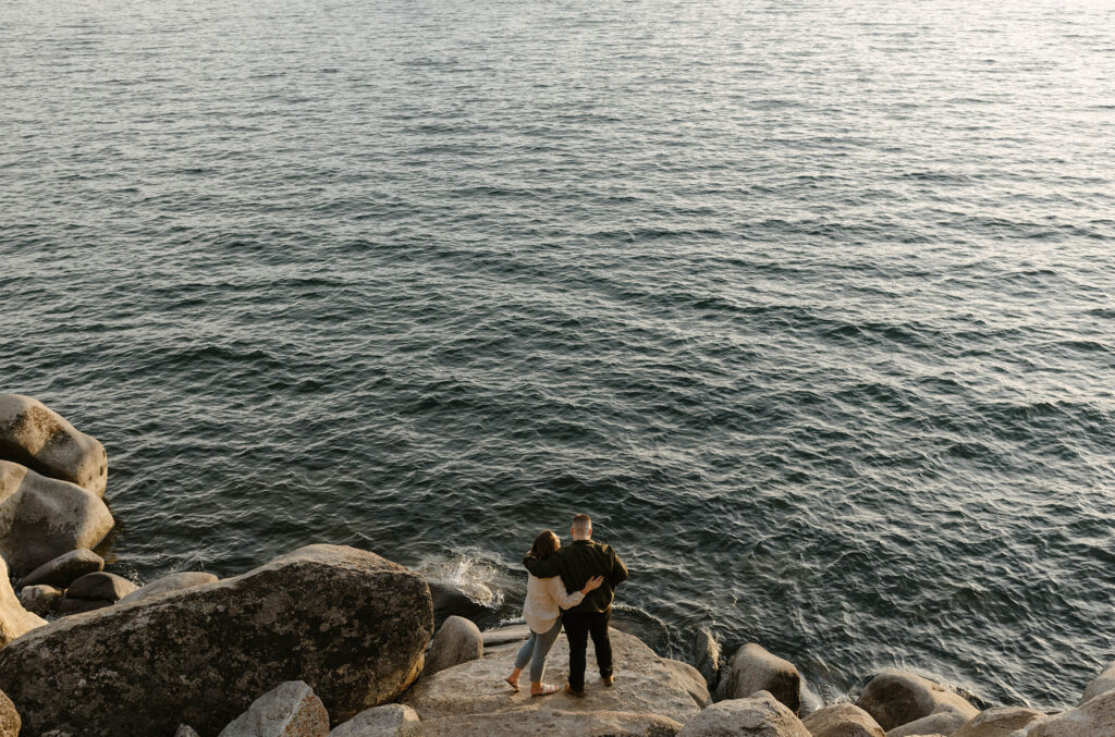Engagement couple with arms around each other standing on large rock next to Lake Tahoe looking out at water at Logan Shoals